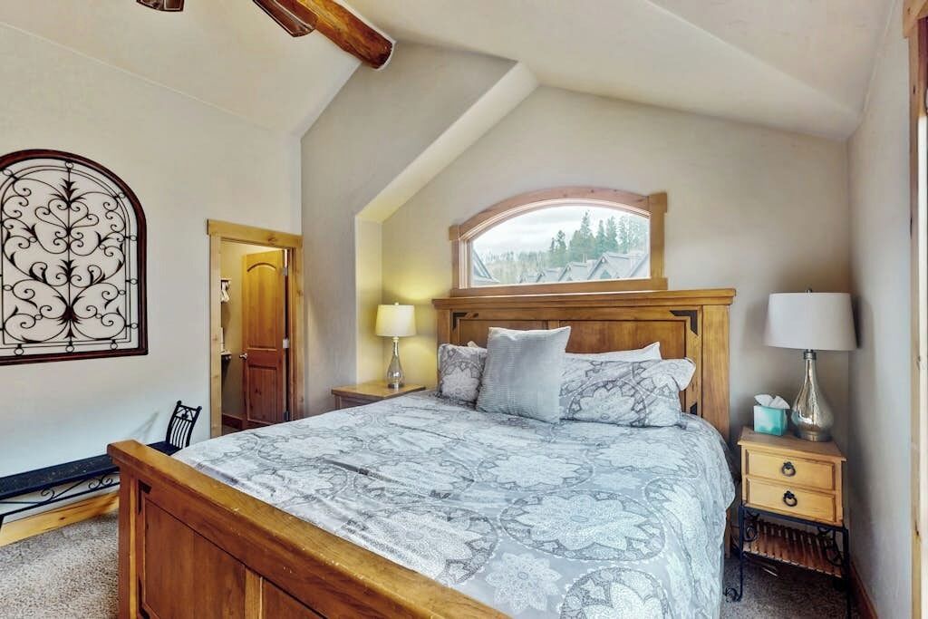 A bedroom with a sloped ceiling, a wooden bed, a nightstand with a lamp, and a window with a view of trees.