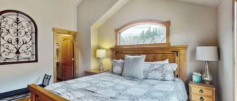 A bedroom with a sloped ceiling, a wooden bed, a nightstand with a lamp, and a window with a view of trees.