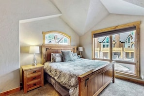 Ein Schlafzimmer mit einem hölzernen Bett, zwei Nachttischen, einem Fenster mit Blick auf Häuser und einer geneigten Decke.