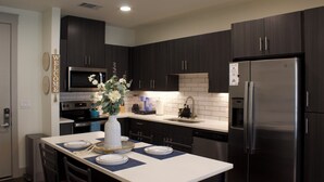A modern kitchen with dark cabinetry, stainless steel appliances, and a central island with seating. - King Bed + Queen Bed - In the Heart of Addison’s Social Scene (Addison)