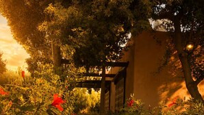 Ein gepflasterter Weg, gesäumt von üppigem Grün und roten Blüten, führt zu einer Holzhütte mit Pergola.