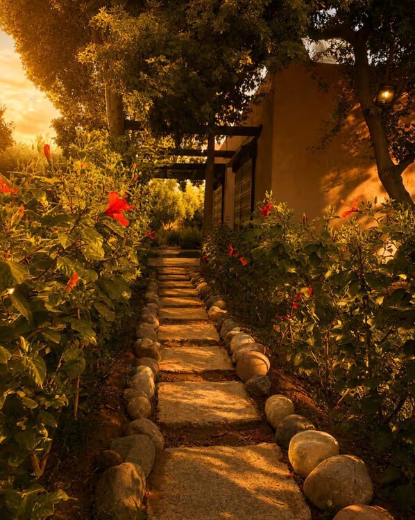 Un sendero de piedra bordeado de exuberante vegetación y flores rojas, que conduce a una estructura de pérgola de madera.