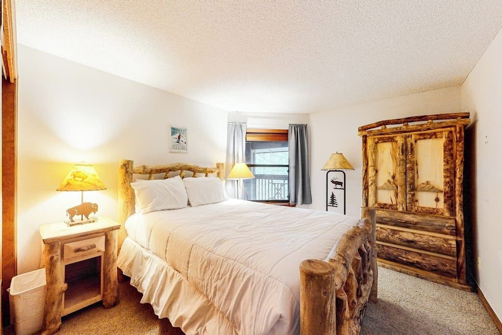 Ein Schlafzimmer mit einem hölzernen Bett, zwei Nachttischlampen, einem Holzschrank, einem Fenster mit Vorhängen und einem kleinen Bild an der Wand.
