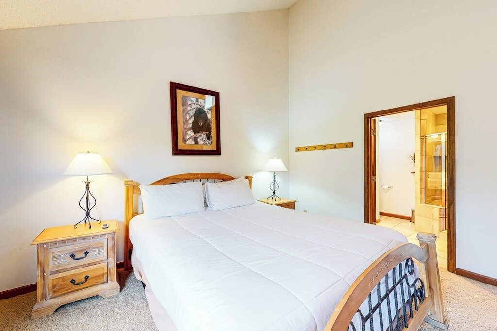 Une chambre avec un grand lit, deux tables de chevet avec des lampes, un tableau encadré au mur et un miroir sur la porte.