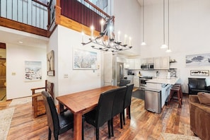 A spacious living area with a wooden dining table, black chairs, a chandelier, and a kitchen in the background.