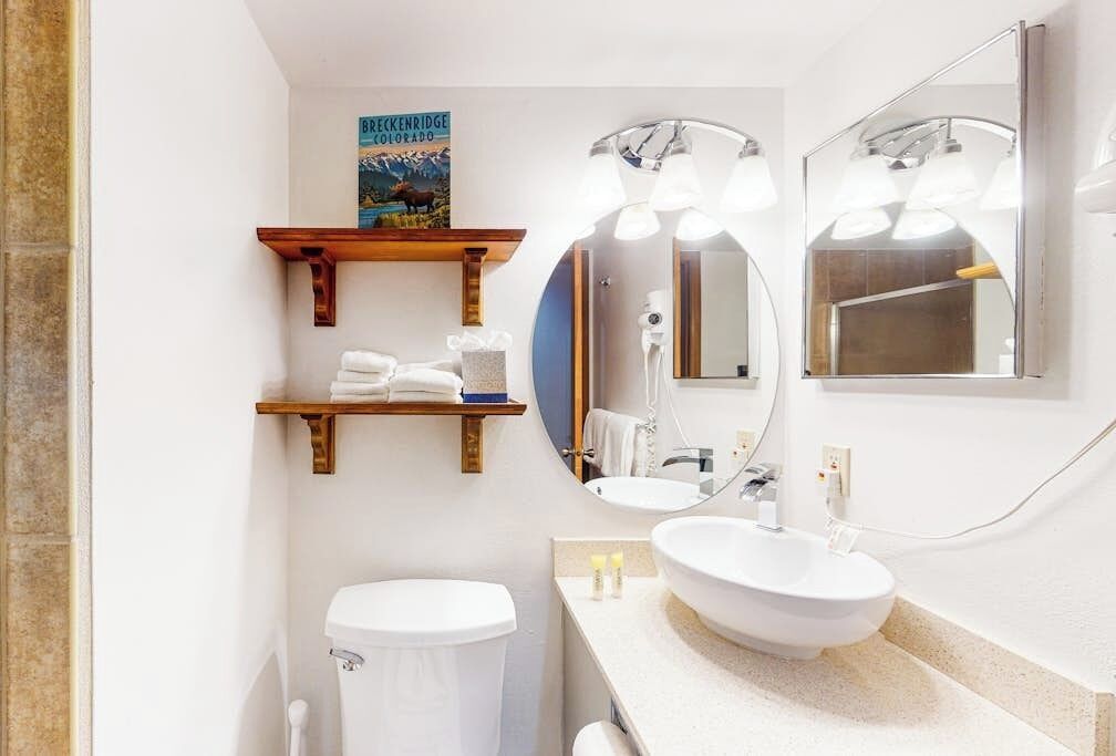A bathroom with a toilet, a sink, a mirror, and a shelf with towels.