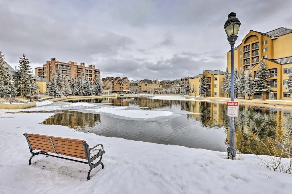 A snow-covered outdoor area with a bench, a lamp post, and buildings in the background.