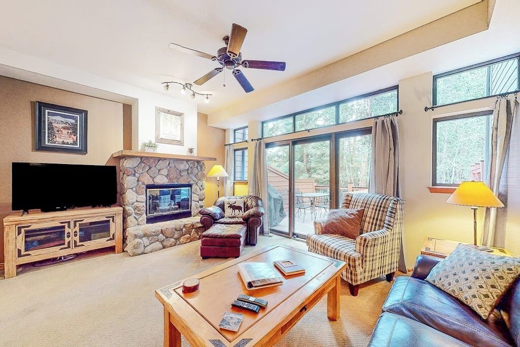 Un salon avec une cheminée en pierre, un téléviseur, un ventilateur de plafond et une grande fenêtre avec des rideaux.