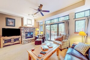 Ein Wohnzimmer mit Kamin aus Stein, einem Fernseher, einem Deckenventilator und einem großen Fenster mit Vorhängen.