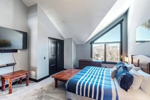 Ein Schlafzimmer mit Bett, Fernseher, einer Holzbank, einem Schminktisch und einem großen Fenster.