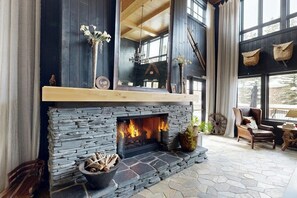 Ein Kamin aus Stein mit Feuer, eine Holzbank und ein Sessel in einem Raum mit großen Fenstern und hoher Decke.
