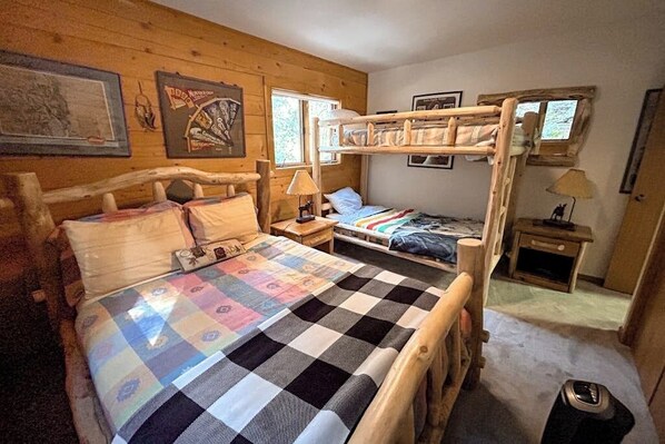 A wooden cabin bedroom with bunk beds, a quilted bedspread, a nightstand, a lamp, and framed pictures on the wall.