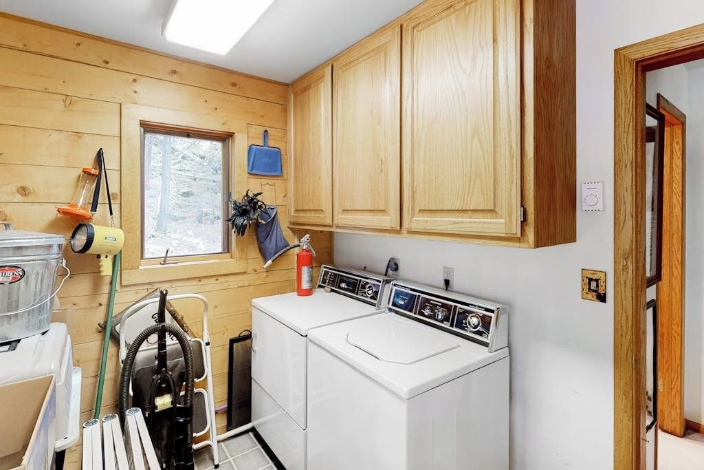 A laundry room with wooden cabinets, a washing machine, a dryer, and a window with a view of trees.