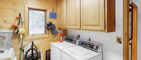 A laundry room with wooden cabinets, a washing machine, a dryer, and a window with a view of trees.
