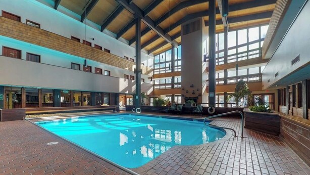 An indoor swimming pool with a brick floor, surrounded by a multi-level building with large windows and a high ceiling. - 2nd-floor mountain-view condo with sauna, balcony, kitchen - walk to lifts (Crested Butte)