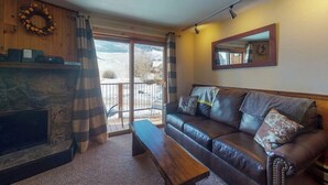 A living room with a fireplace, a leather sofa, a bench, and a balcony with a view. - 2nd-floor mountain-view condo with sauna, balcony, kitchen - walk to lifts (Crested Butte)