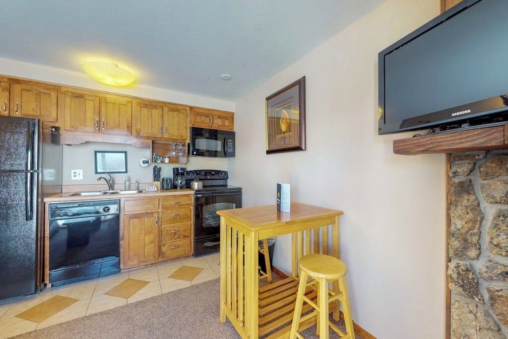 A compact kitchen with wooden cabinets, a microwave, a stove, and a dishwasher. There's a small wooden table with a stool, a flat-screen TV mounted on a stone wall, and a framed picture on the wall.