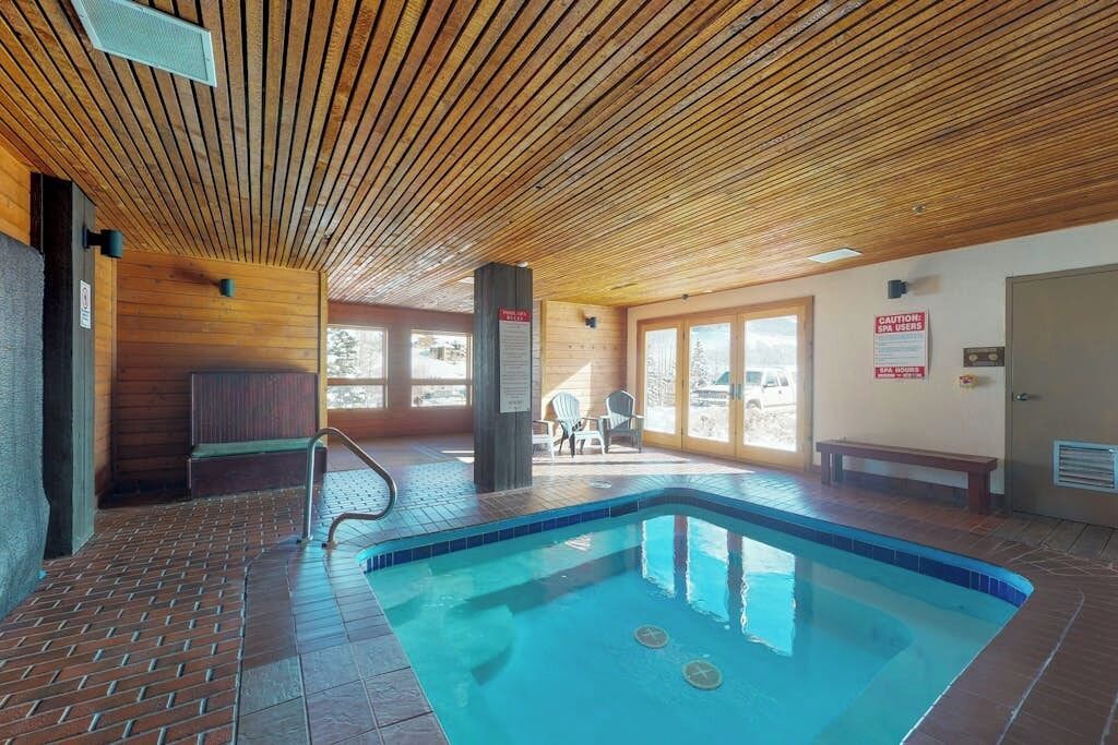 An indoor swimming pool with wooden ceiling, tiled floor, and a bench near the pool.