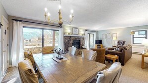 A dining area with a large wooden table, cushioned chairs, a chandelier, and a stone fireplace. - Mountain-view condo with private deck, fireplace & hot tub (Crested Butte)