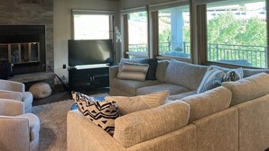 A living room with a sofa, a flat-screen TV, a fireplace, and large windows. - Rustic retreat with slopeside access, mountain views & cozy fireplace (Crested Butte)