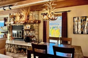 A stone fireplace with a rustic decor, a dining table with chairs, and a view of the outdoors through a window. - Mountain view condo with fireplace & W/D and access to shared hot tub (Crested Butte)