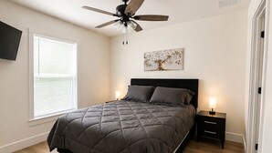 Ein Schlafzimmer mit einem Bett, einem Deckenventilator, einem Nachttisch, einem an der Wand montierten Fernseher und einem Fenster mit Jalousien.