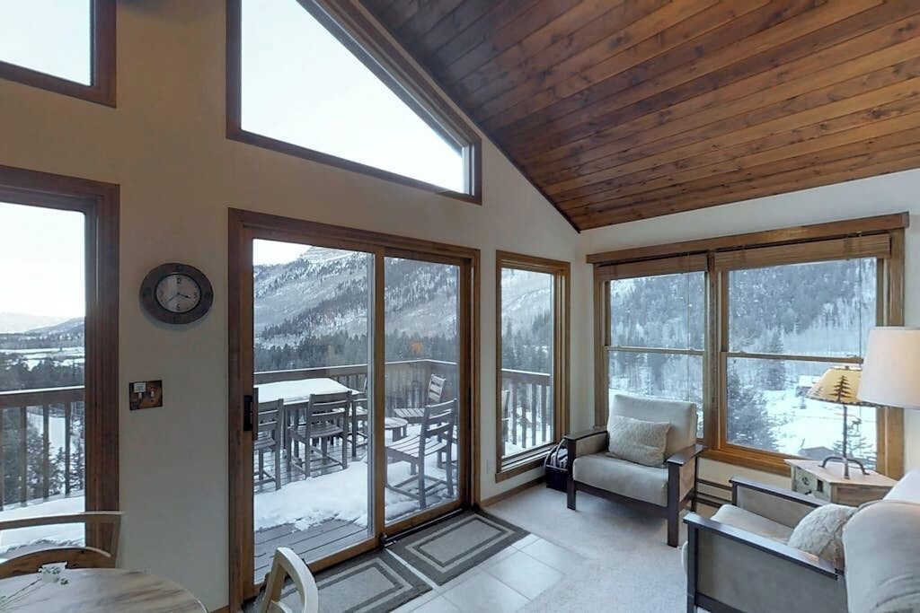A cozy room with a wooden ceiling, large windows, a clock, and a view of a snowy outdoor deck.