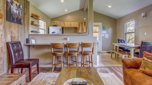 A living room with a wooden dining table, four chairs, a brown leather sofa, and a kitchen area with wooden cabinets and a refrigerator.