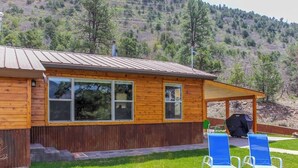 A wooden cabin with a covered patio, a grill, and two lounge chairs.