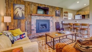 A living room with a fireplace, wooden panel walls, a sofa, a coffee table, and a kitchen area with chairs and a refrigerator.
