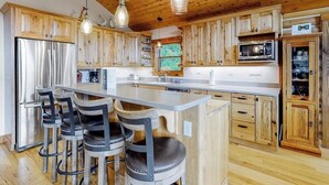 A kitchen with wooden cabinets, a stainless steel refrigerator, a microwave, and a bar area with stools. - Secluded home with amazing mountain & lake views, deck, fireplace, & WiFi (Durango)