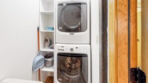 Pieni ja käytännöllinen pyykinhuone, jossa on kaksi päällekkäin asetettua pesukonetta, hylly, jossa on sininen pyyhe, ja puinen ovi.