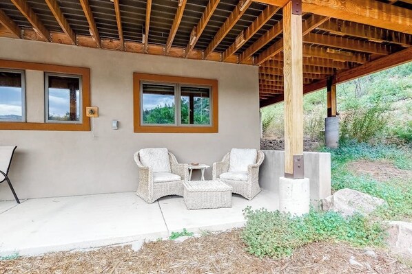 Ein überdachter Patio-Bereich mit zwei Korbstühlen und einem kleinen Tisch, darüber Holzträger und ein Blick ins Grüne.