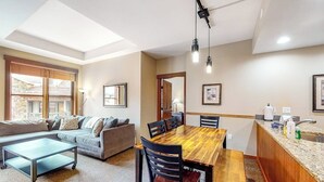 A modern living room with a sofa, a wooden dining table, and a kitchen area with a sink and countertop. - Ski-In/Ski-Out Modern Condo with Shared Hot Tub & Sauna - Views of the Village (Copper Mountain)