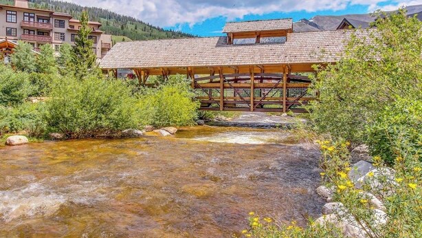 A lodge with a wooden deck overlooking a clear stream, surrounded by greenery and mountains. - Ski-In/Ski-Out Modern Condo with Shared Hot Tub & Sauna - Views of the Village (Copper Mountain)