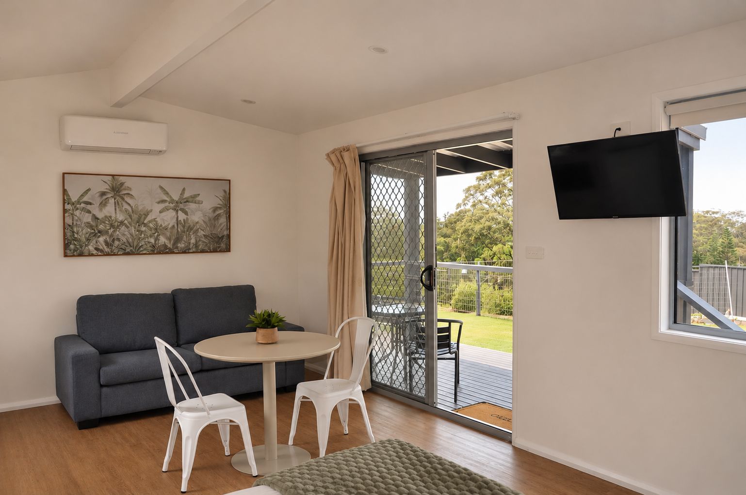A modern living room with a grey sofa, a round table with chairs, a potted plant, and a sliding glass door leading to an outdoor area.