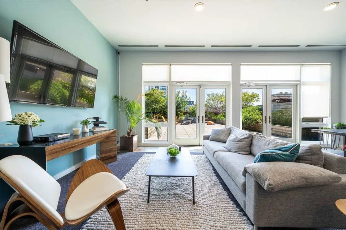 A modern living room with a sofa, coffee table, and chairs, a desk with a computer, and large windows with a view of greenery.