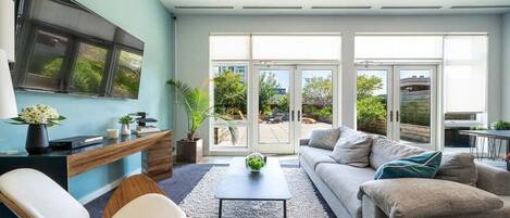 A modern living room with a sofa, coffee table, and chairs, a desk with a computer, and large windows with a view of greenery.