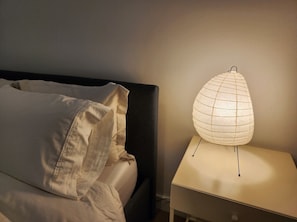 A bed with pillows, a bedside table with a lamp, and a wall. - Hoboken Furnished Housing – Modern 1BR with Balcony, Elevator, W/D, Mins to NYC (Hoboken)