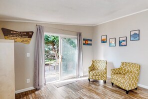 Ein Zimmer mit einer Schiebeglastür, die zu einem Balkon führt, zwei gemusterte Sessel und eine Wand mit gerahmten Kunstwerken.