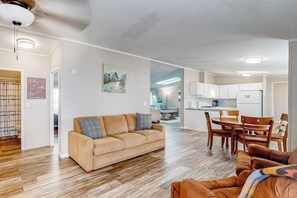 Un salon avec un canapé, un coin repas avec une table et des chaises, une cuisine équipée de placards et d’un réfrigérateur, et un ventilateur de plafond.