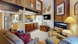 Una sala de estar acogedora con un sofá, una mesa de centro y un televisor. Hay una zona de cocina con barra y taburetes.
