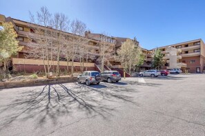Ein Parkplatz mit mehreren Autos, ein Gebäude mit mehreren Stockwerken und Balkonen, sowie Bäume im Vordergrund.