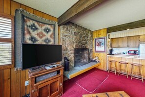 Ein Wohnzimmer mit Kamin aus Stein, Holzwänden, einem Fernseher und einer Küchenzeile mit Barhocker.