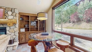 Een gezellige kamer met een stenen openhaard, houten planken, een tafel met glazen blad en een groot raam met uitzicht op bomen.