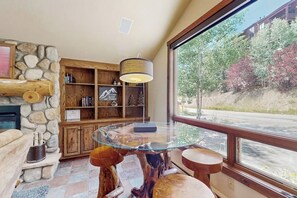 Ein gemütliches Zimmer mit Kamin aus Stein, Holzregalen, einem Tisch mit Glasplatte und einem großen Fenster mit Blick auf Bäume.