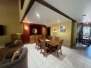 Uma área de refeições com mesa e cadeiras de madeira, uma cozinha ao fundo e uma escada que leva a um andar superior.