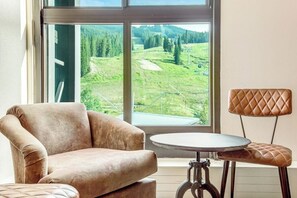 Ein Zimmer mit einem großen Fenster, einem gepolsterten Sessel, einem kleinen runden Tisch und einem Stuhl mit einer gesteppten Rückenlehne.