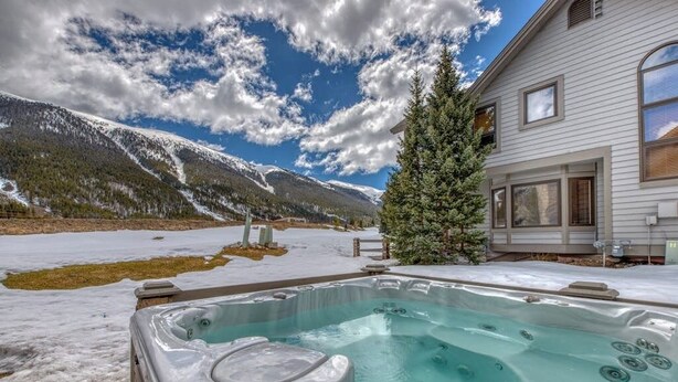 A hot tub with a mountainous backdrop, a wooden deck, and a house with large windows. - Spacious resort home w/private garage, hot tub & laundry. Bus to slope (Copper Mountain)