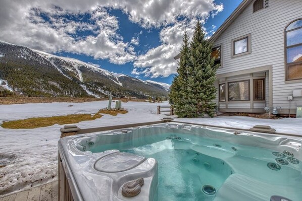 Ein Whirlpool vor einer Bergkulisse, eine Holzterrasse und ein Haus mit großen Fenstern.